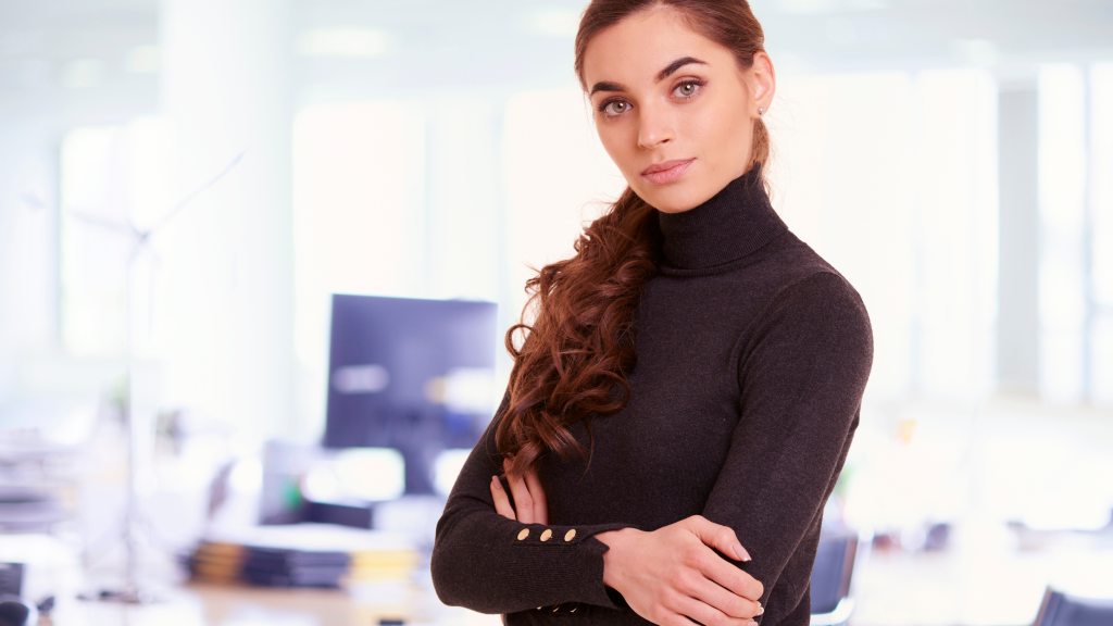 Professional woman with long braided hair and a dark turtleneck stands with arms crossed in a bright office setting, looking at the camera.