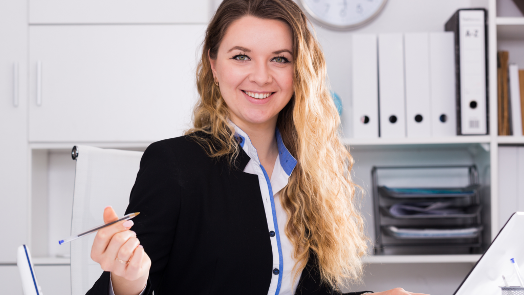 Smiling woman in a black blazer holding a pen at a desk in a bright office with binders on shelves behind her.