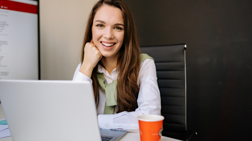 Smiling woman seated at a desk with a laptop, resting her chin on her hand in a modern office setting with a red coffee cup nearby.