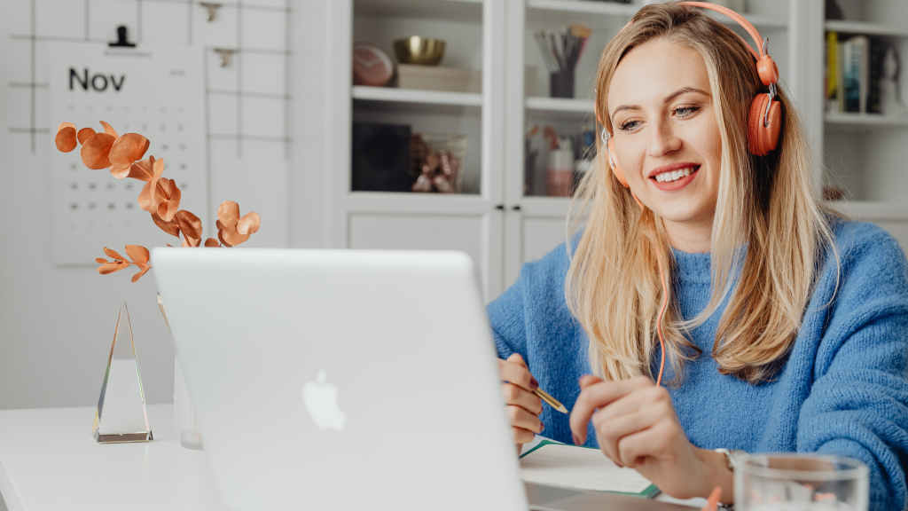 Woman wearing coral over-ear headphones smiles while working on a laptop at a white desk in a bright home office.
